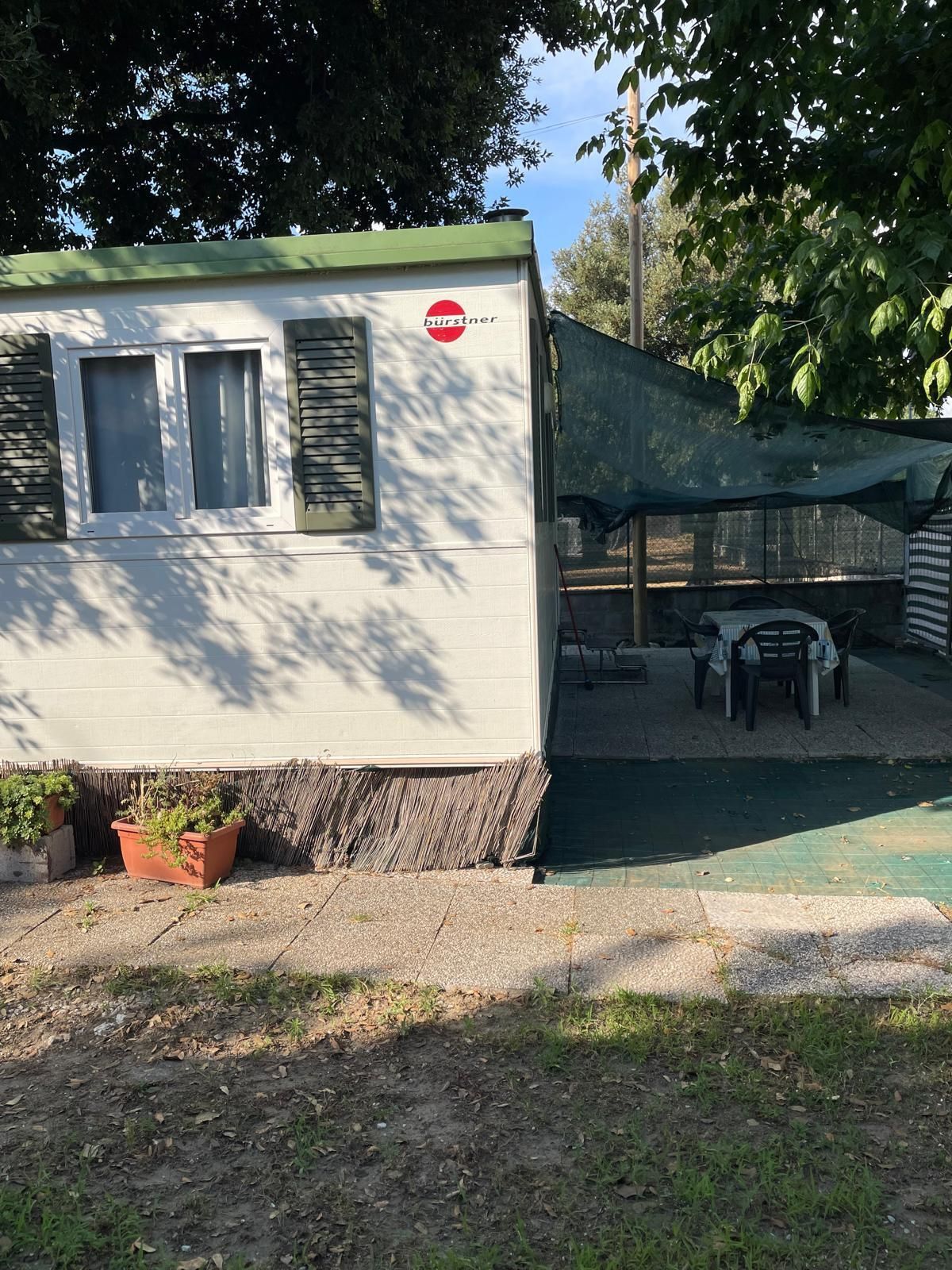 Una casa mobile bianca con tetto verde e finestre con persiane, dotata di un patio esterno ombreggiato con tavolo e sedie.