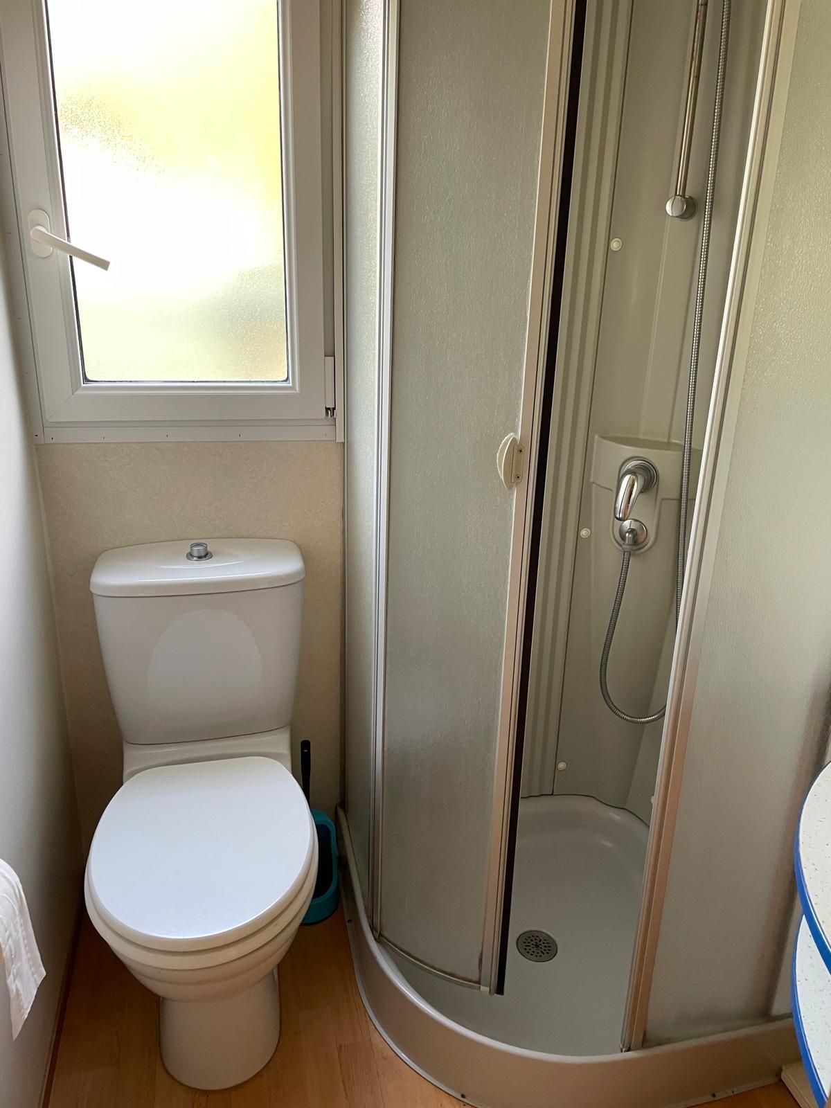 In un bagno, accanto a una piccola cabina doccia angolare chiusa con porte in vetro e una finestra, si trova un WC bianco.