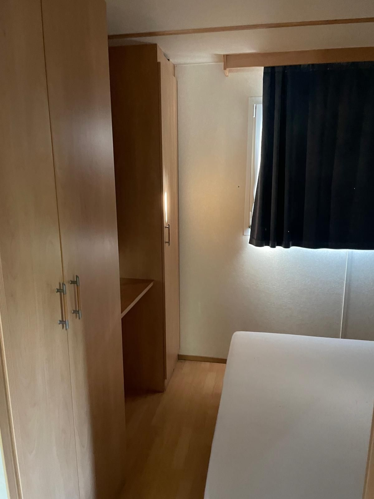 Armadietti in legno chiaro fiancheggiano uno spazio stretto che conduce a una finestra con tende scure, da cui si intravede parzialmente la struttura di un letto bianco.