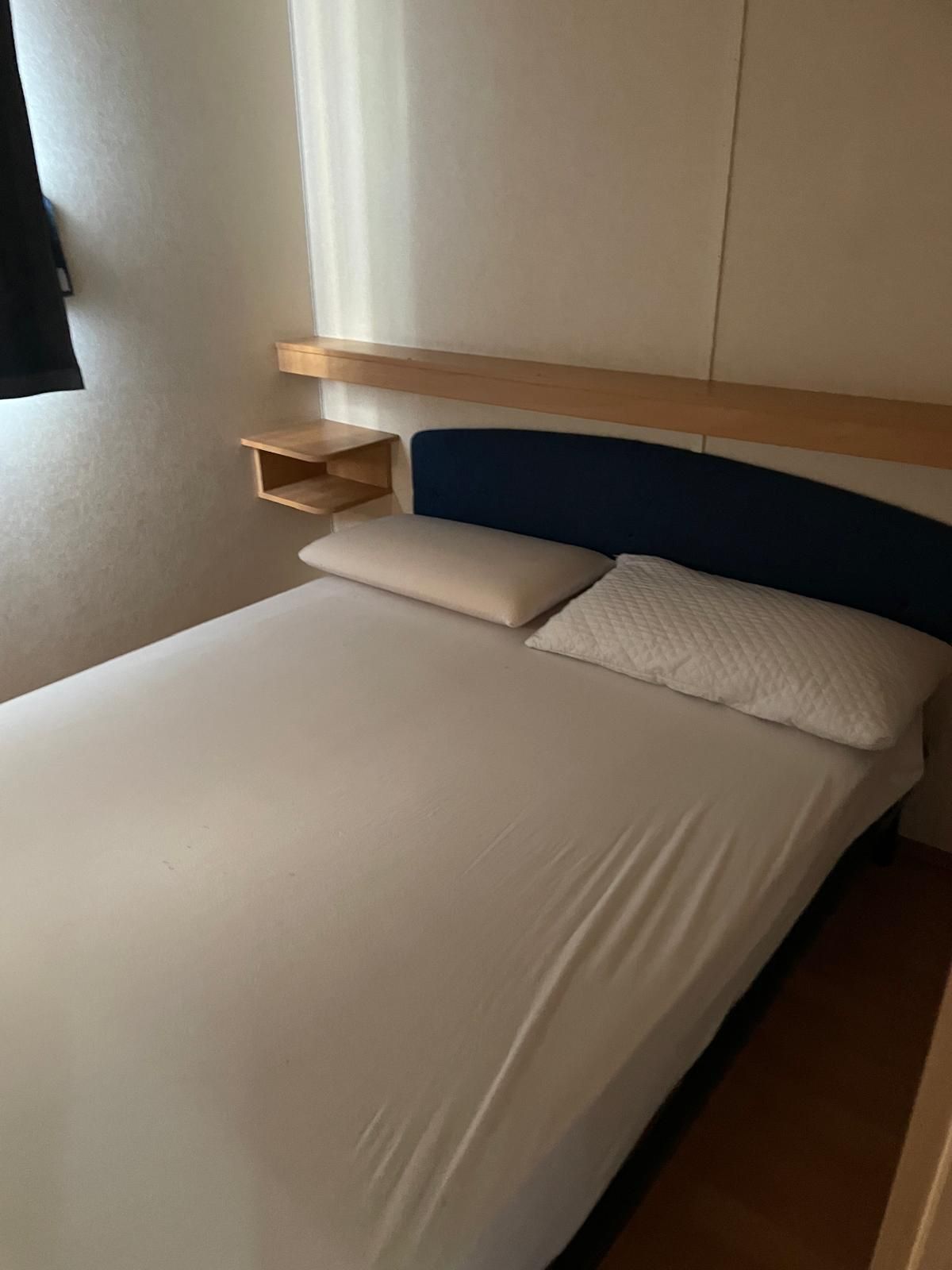 Un letto matrimoniale con lenzuola bianche, una testiera blu scuro e una mensola in legno a muro in una stanza semplice dai toni neutri.