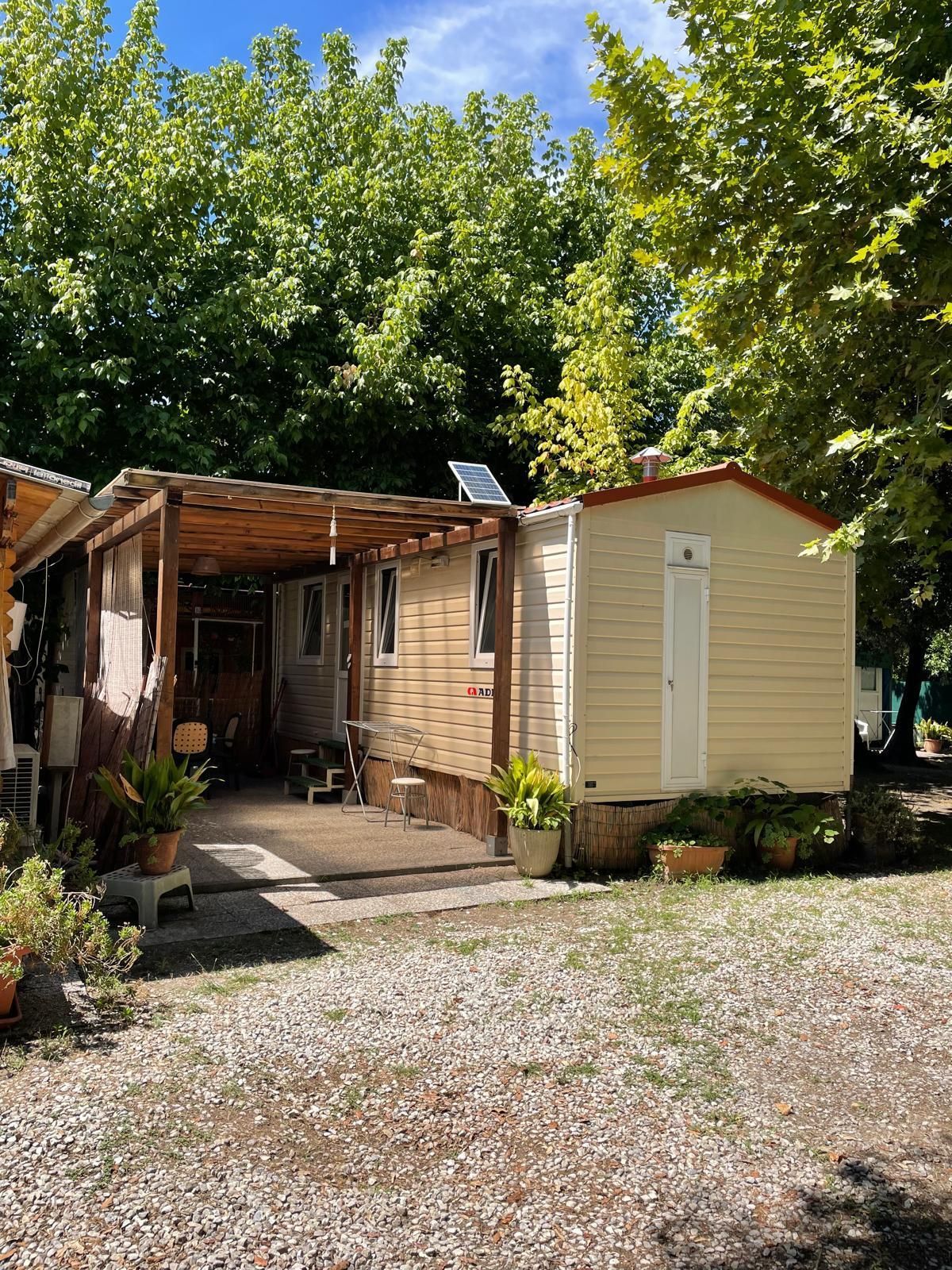 Una casa mobile con patio coperto, immersa in una zona ombreggiata e alberata con pavimento in ghiaia.