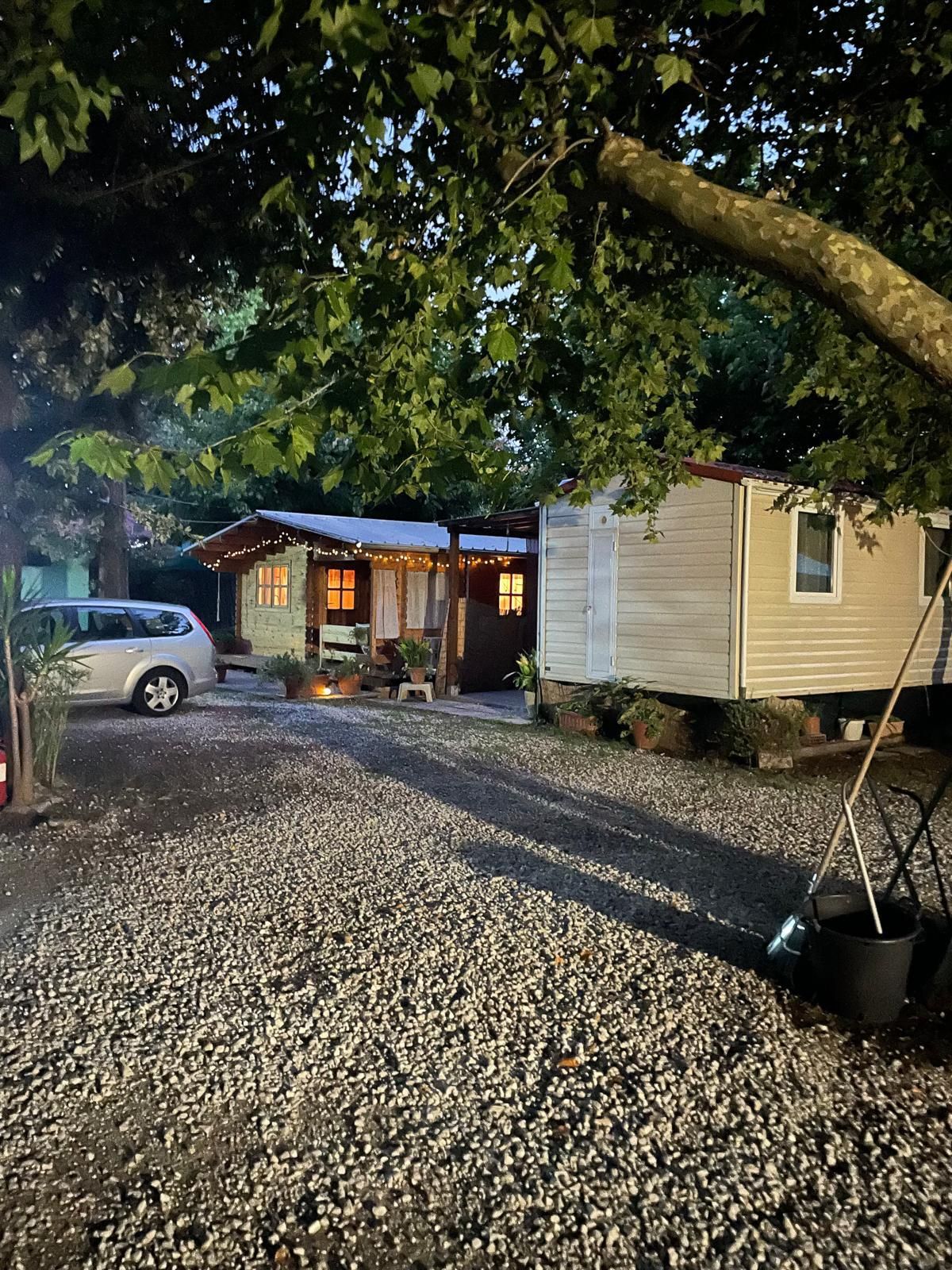 Un'area ghiaiosa con due piccole casette o case mobili illuminate al crepuscolo, con un'auto argentata parcheggiata e degli alberi sullo sfondo.