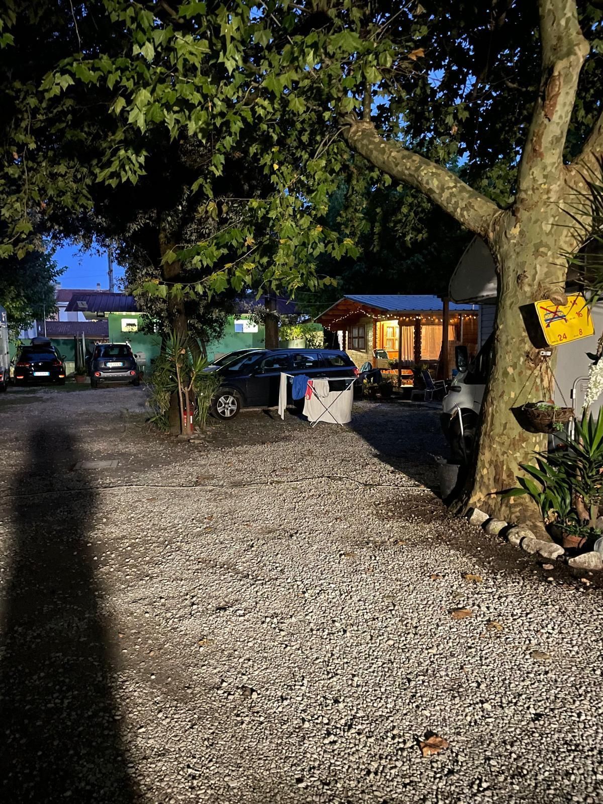 Un parcheggio sterrato al crepuscolo, con auto parcheggiate, un edificio in legno e grandi alberi, con una lunga ombra in primo piano.