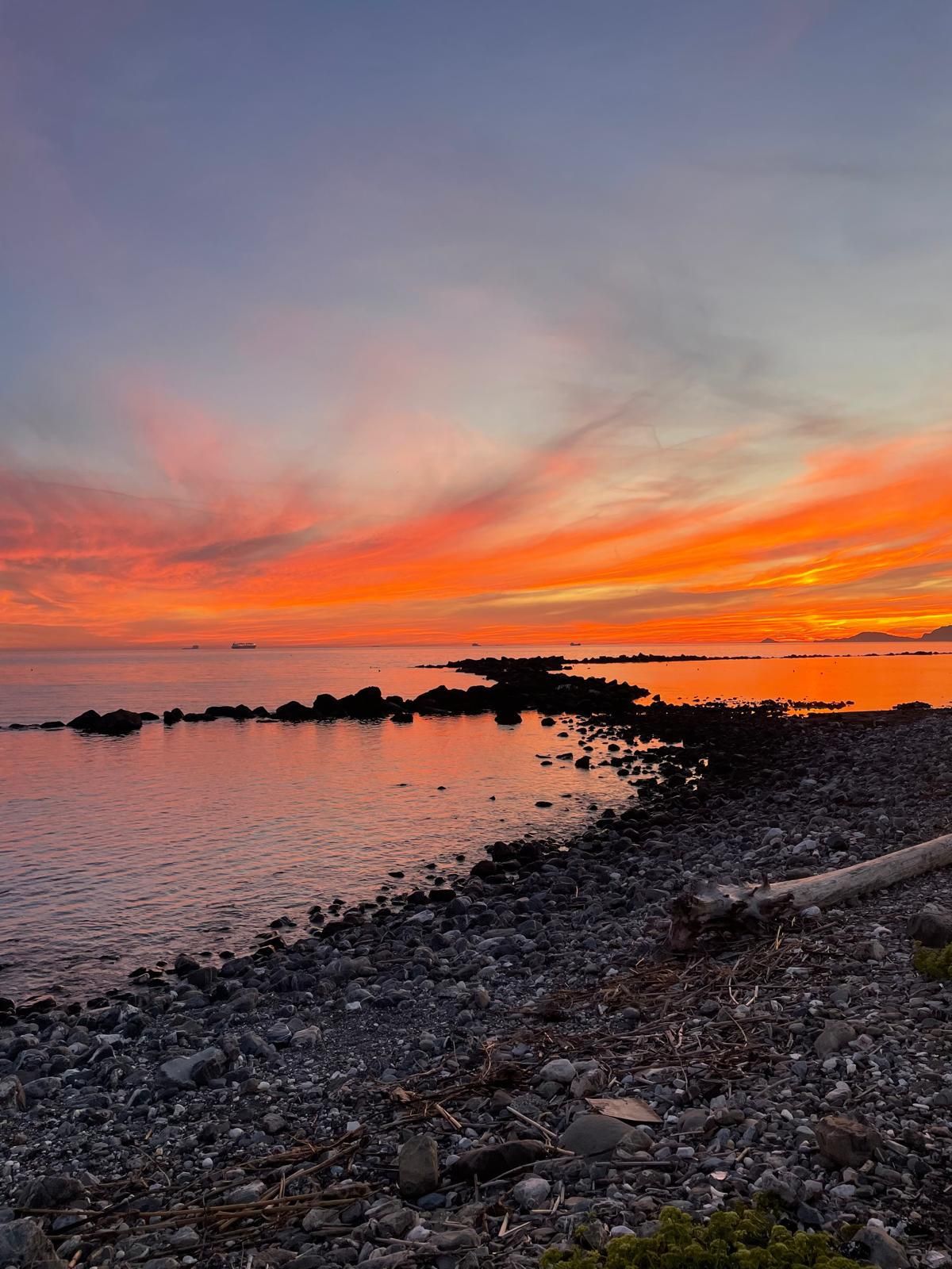 Un tramonto vibrante su una costa rocciosa con un lungo molo di pietra scura che si estende nelle acque calme e riflettenti.