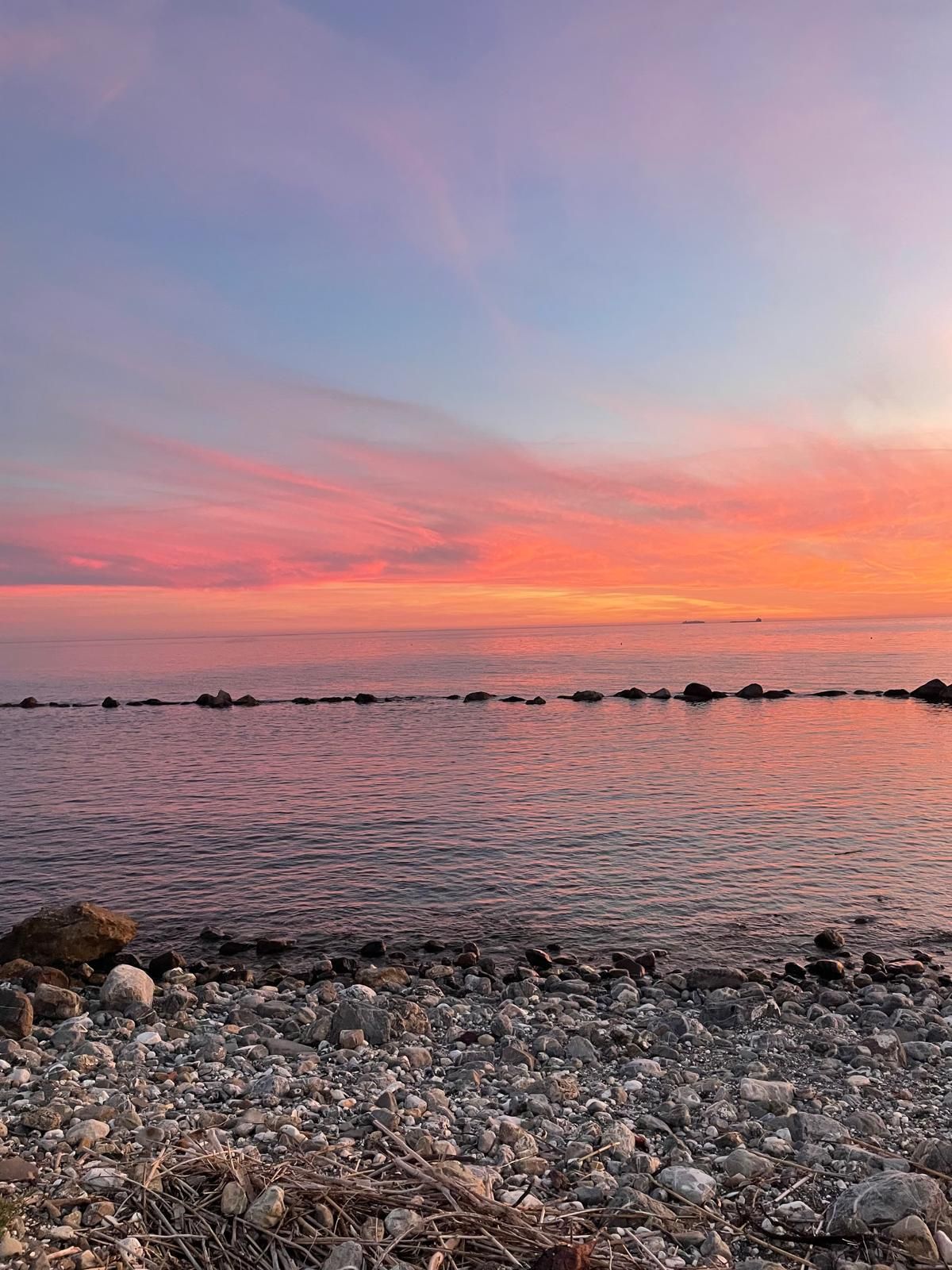 Una vista suggestiva di una spiaggia di ciottoli al tramonto, con nuvole rosa e arancioni che si riflettono sulle calme acque dell'oceano.