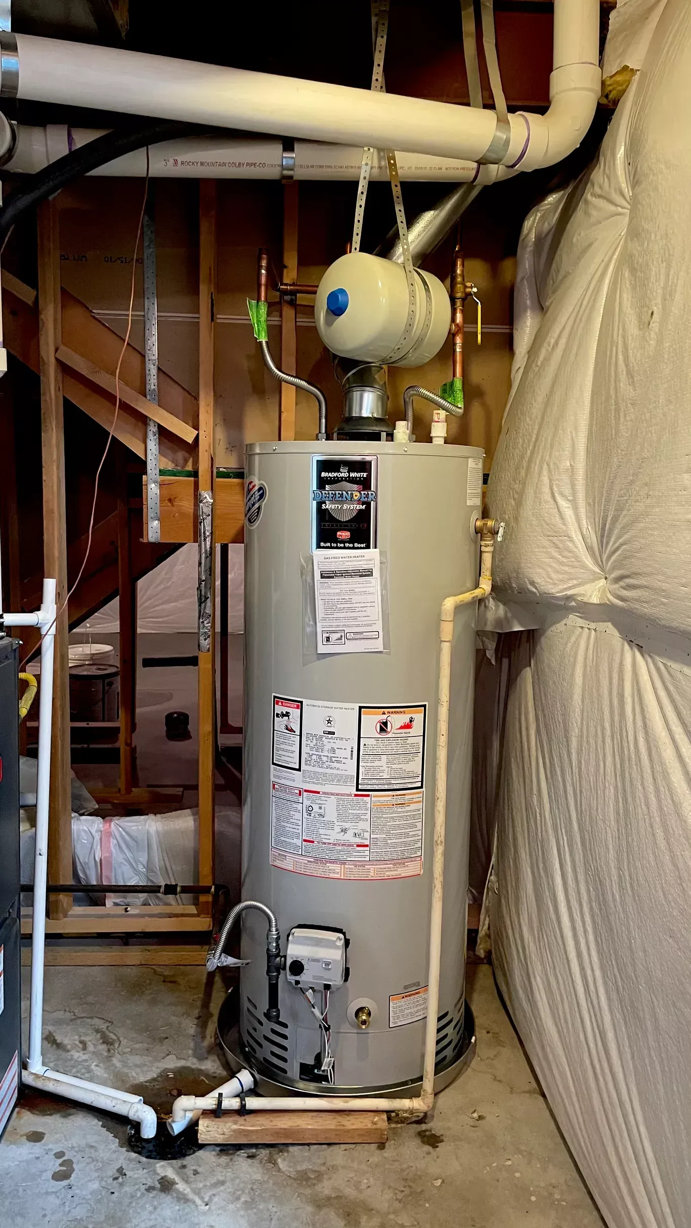 A water heater is sitting in a basement next to a staircase.