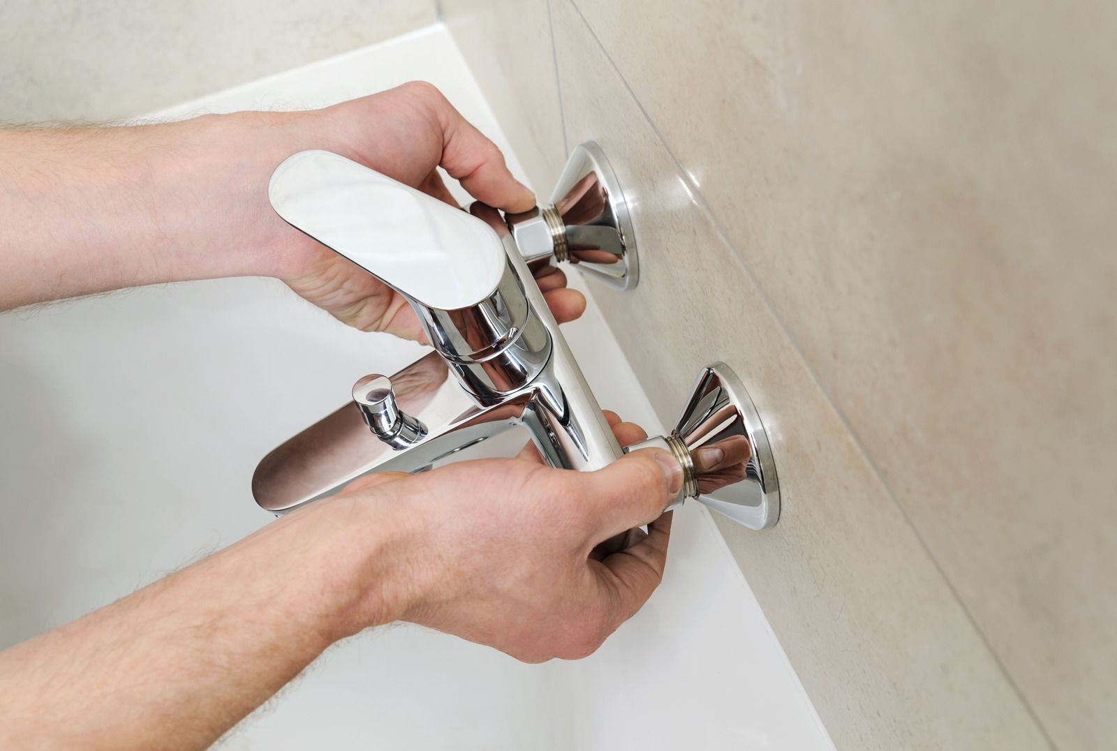 Hands installing a shiny chrome shower faucet on a beige tiled wall.
