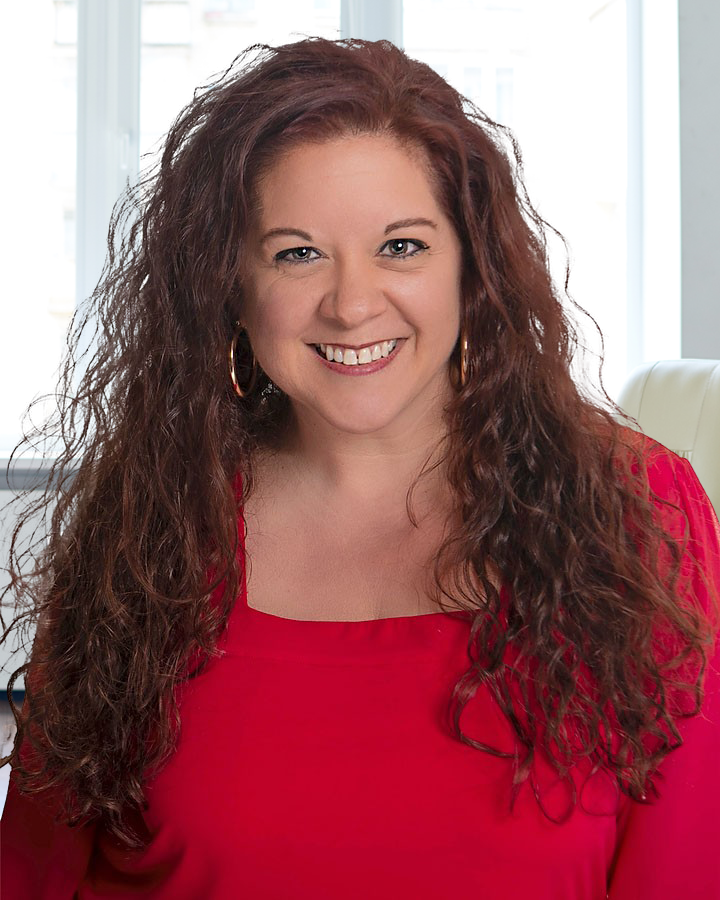 Woman with curly auburn hair smiles, wearing a red top. Indoors, natural light.