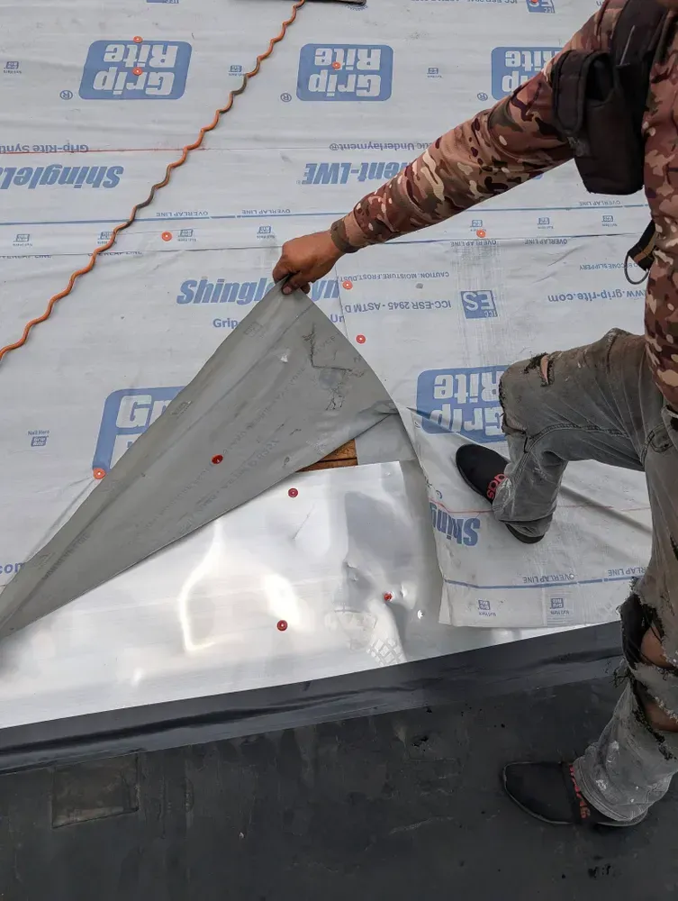 Roofer installing flashing. Holding up a silver metal sheet over a roof surface with blue and white underlayment.