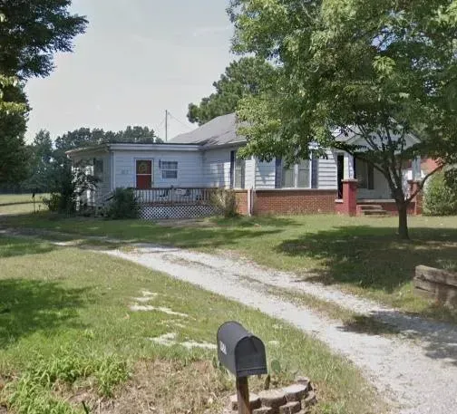 Small, light blue house with porch, gravel driveway, and mailbox in front, trees on the right.