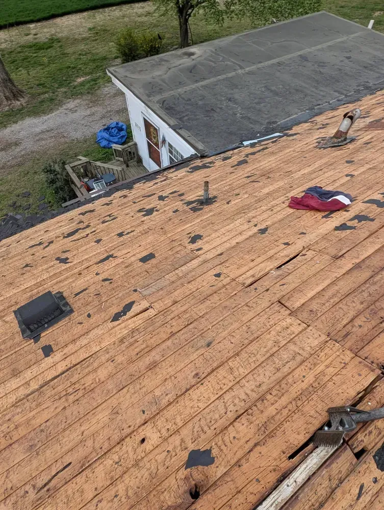 View from a roof showing partially torn shingles and exposed wooden planks, with a small building in the background.