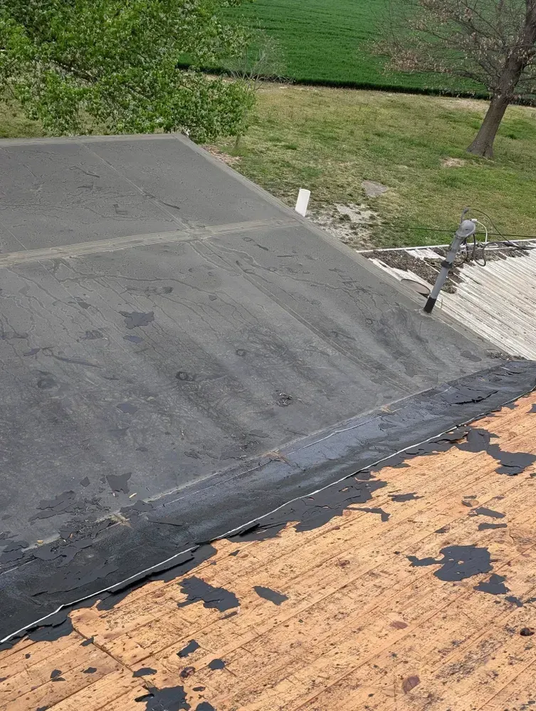 Black flat roof with peeling edges over an older wood-shingled roof, outdoors.