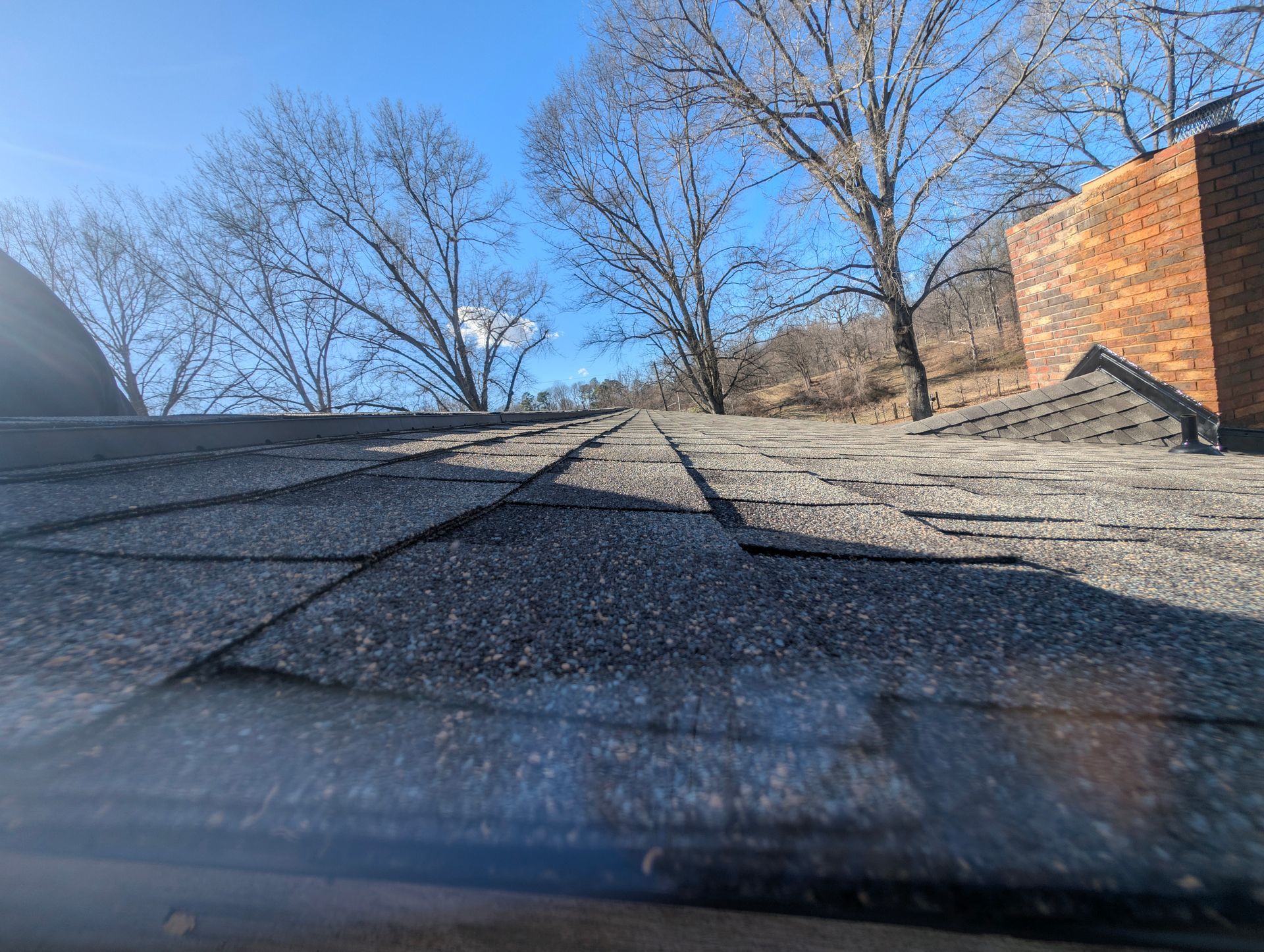 A low-angle view of a weathered asphalt shingle roof, chimney, and bare trees against a blue sky.