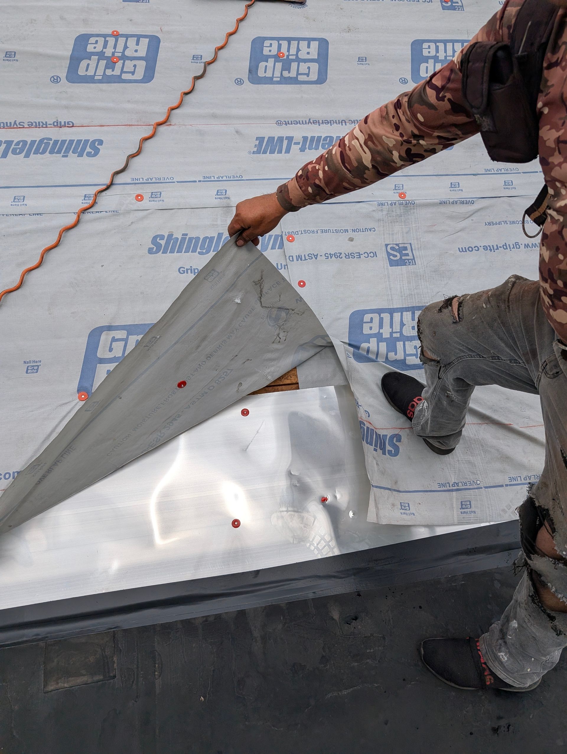 Roofer installing flashing. Metal sheet being lifted, exposing roof membrane. Gray and black.