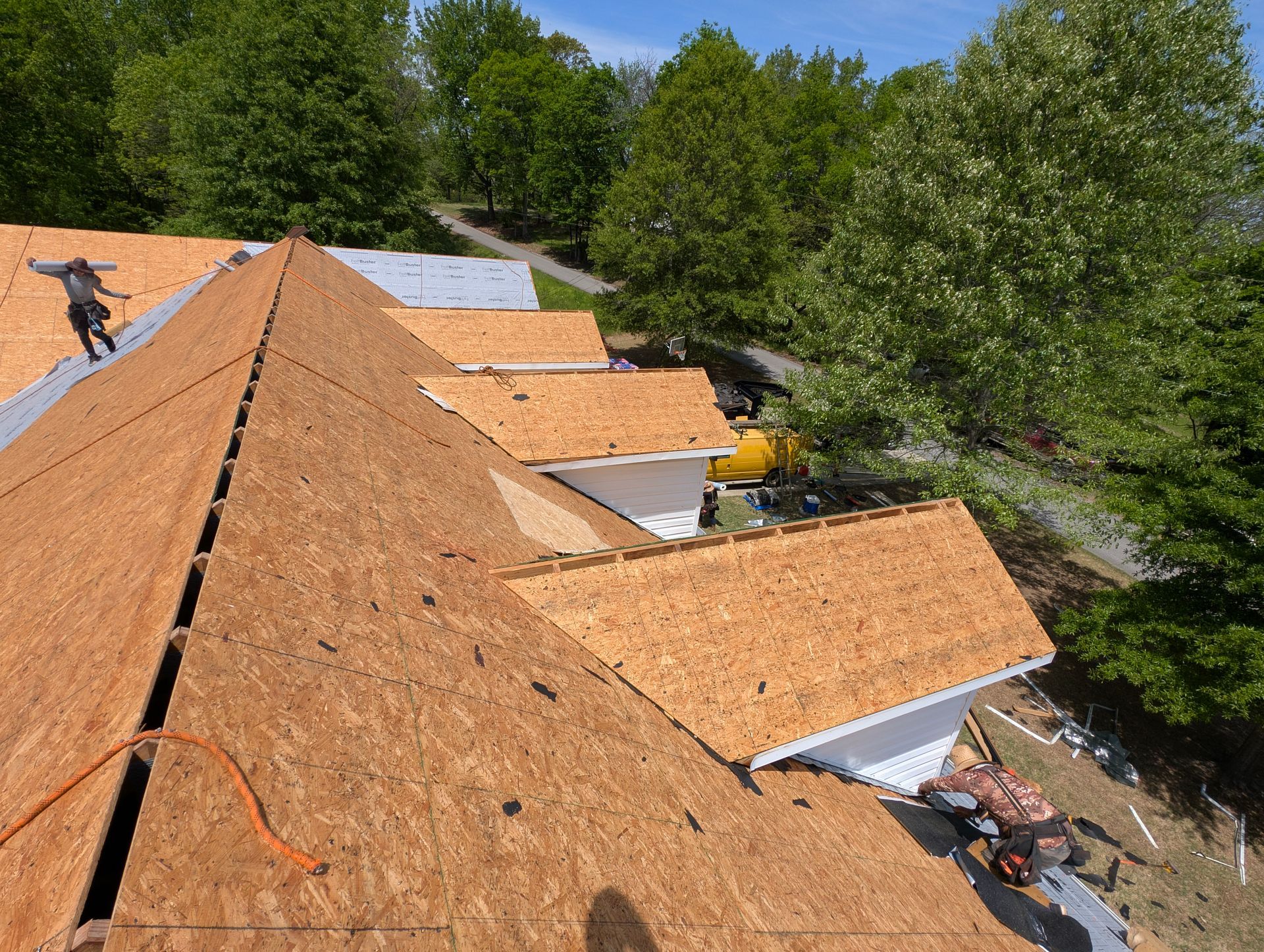 Roofing work in progress; plywood laid on roof; workers; trees in background.
