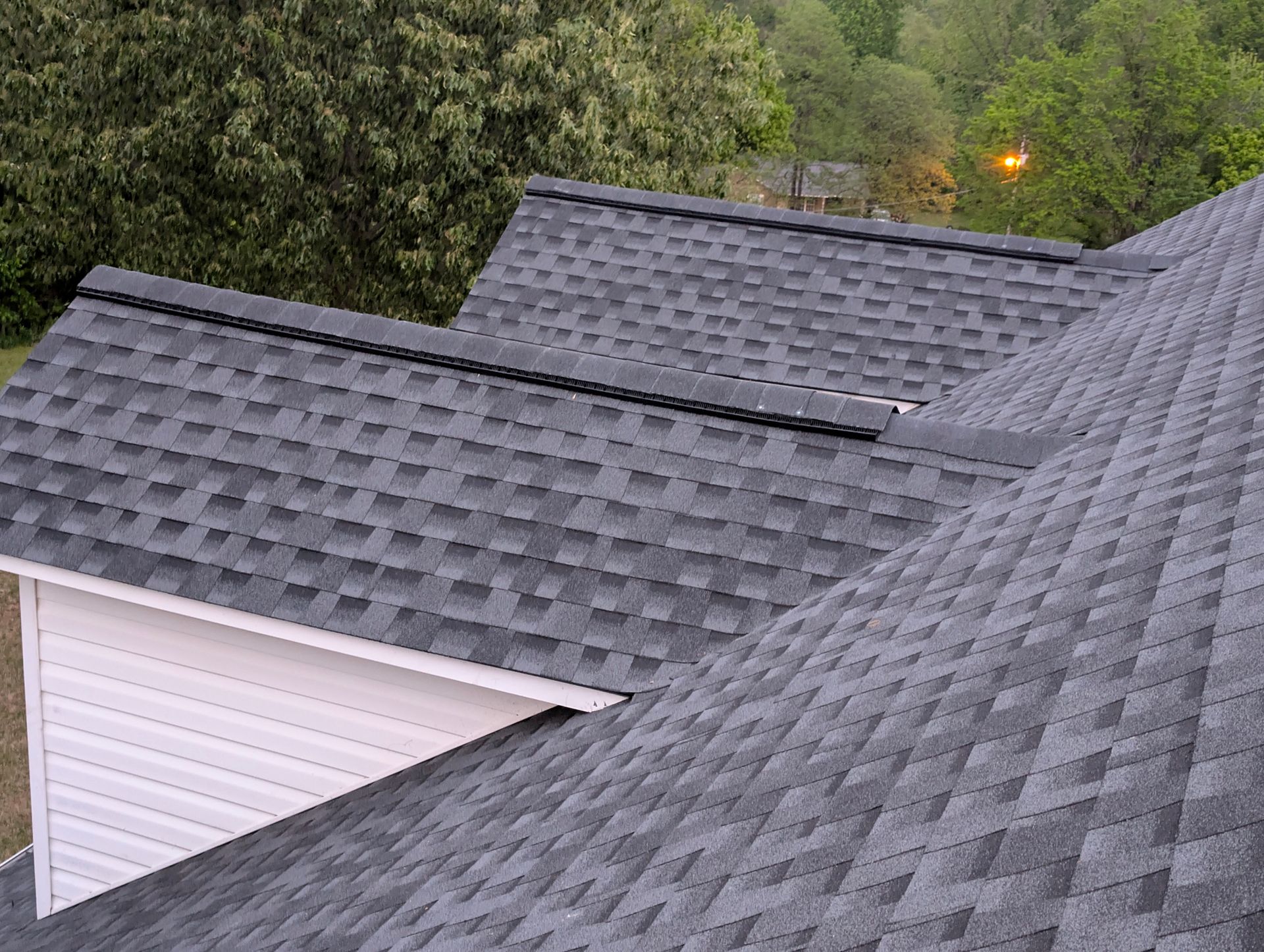 Gray asphalt shingle roof on a house, with a white gable and trees in the background.