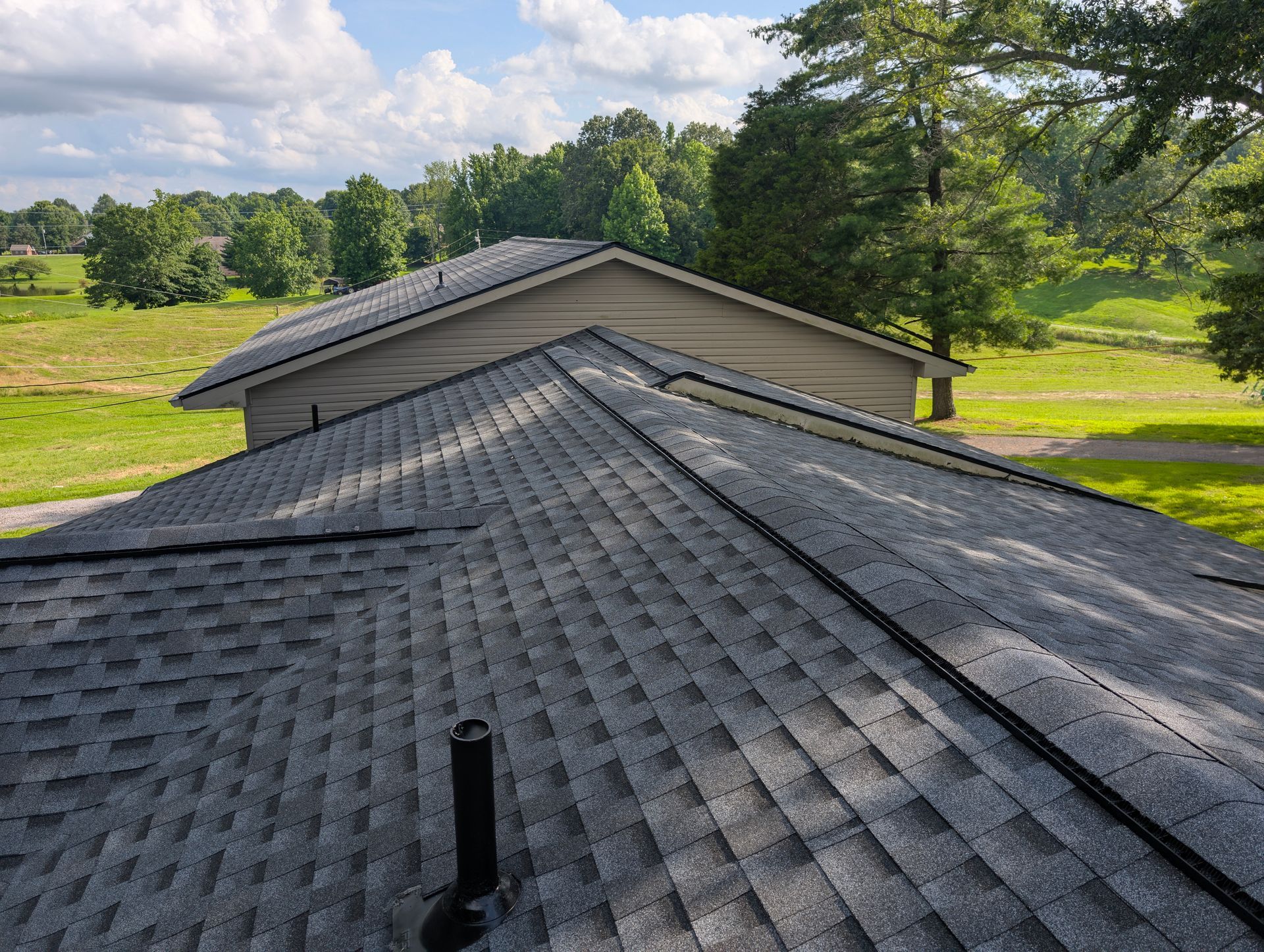 View of two shingled roofs against a blue sky with trees and a green field in the background.