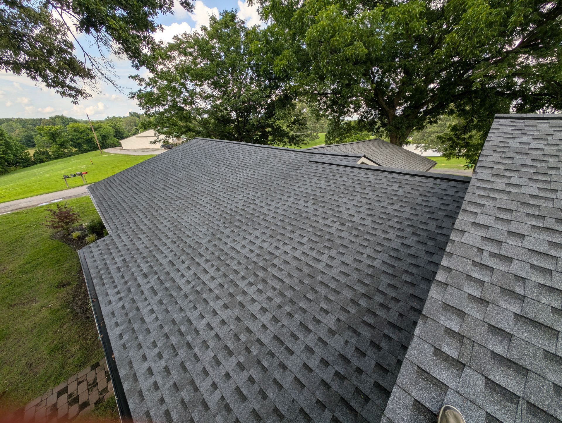 Dark gray asphalt shingle roof on a house, angled view, with trees and grass in the background.