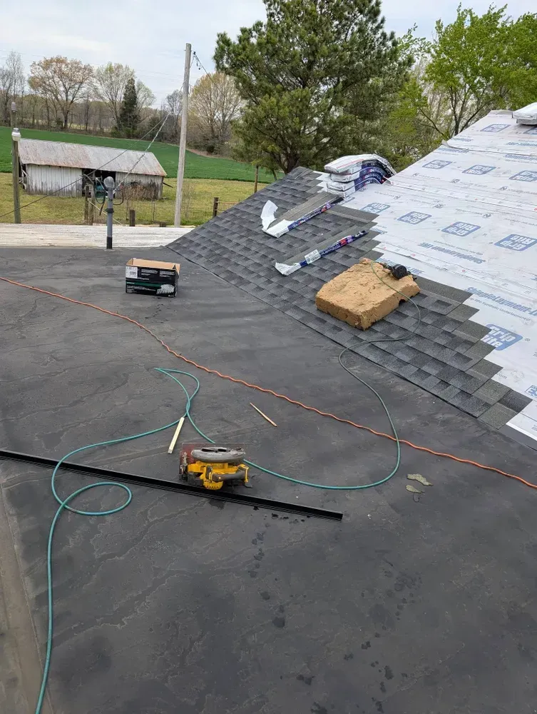 Roof with asphalt shingles being installed, tools visible. Background includes a barn and trees.