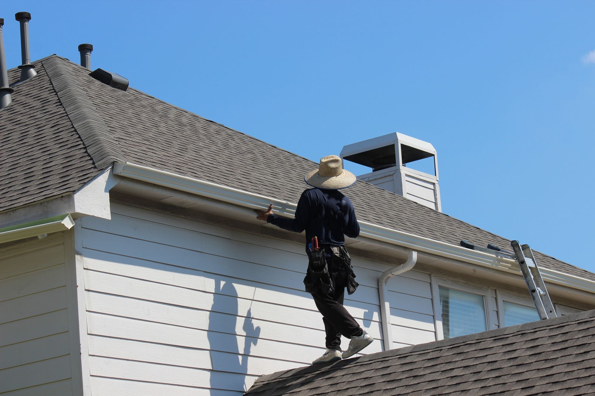 Person in a hat on a roof, inspecting gutters; white siding, brown roof, blue sky.