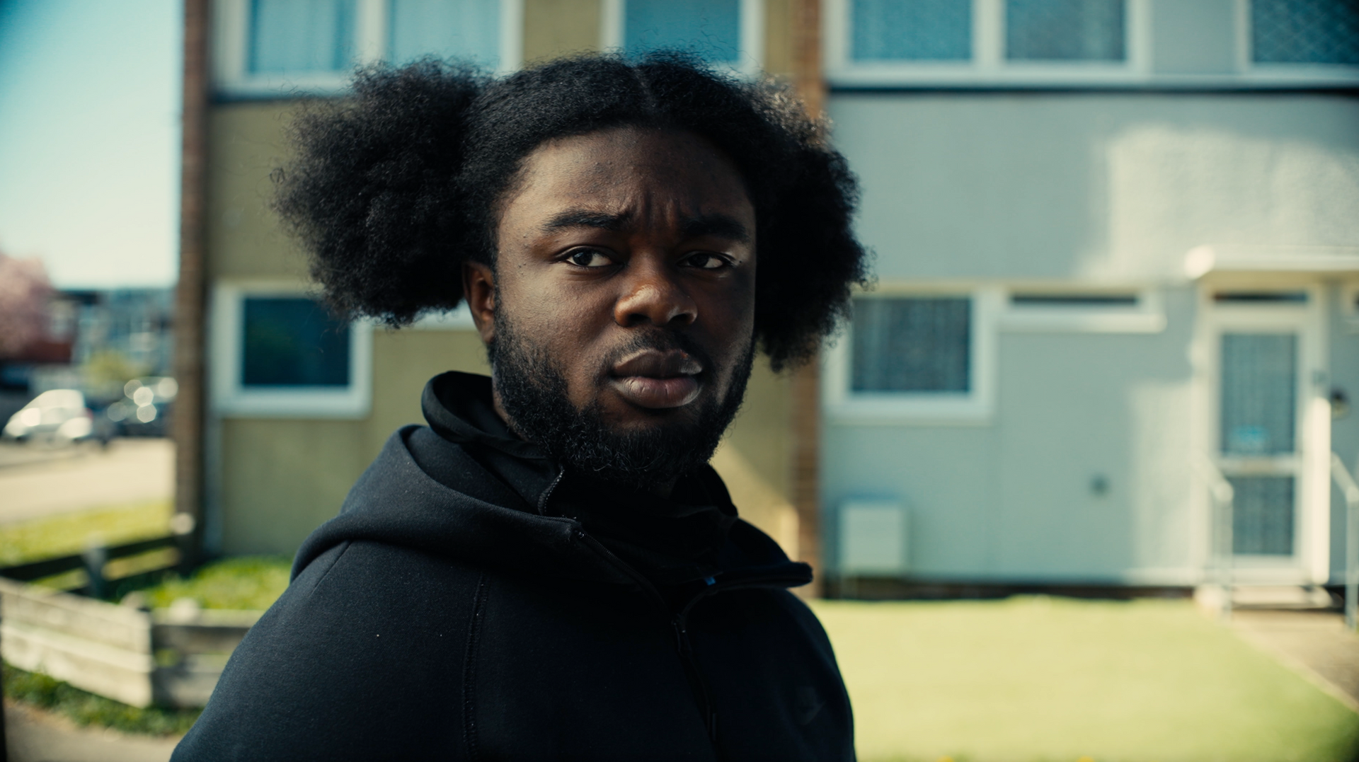A man with curly hair and a beard is standing in front of a building.