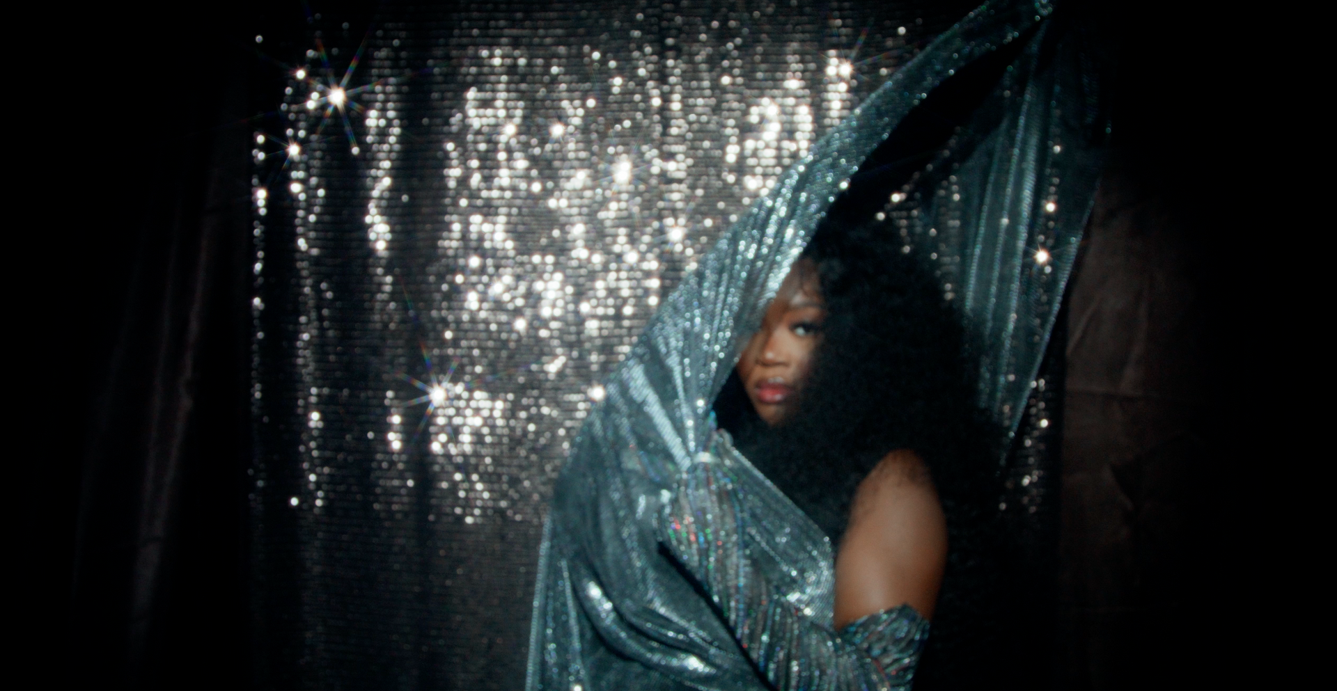 A woman in a blue dress is standing in front of a sequined curtain.
