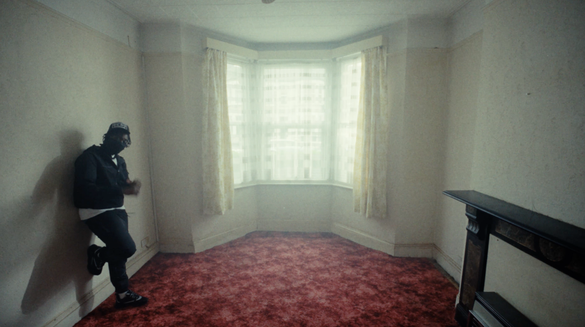 A man in a mask is leaning against a wall in an empty room.