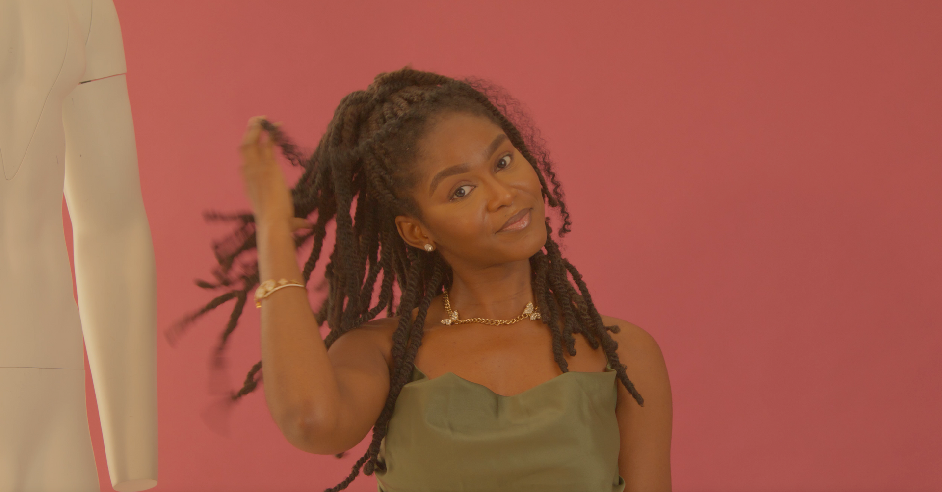 A woman with dreadlocks is standing in front of a pink background.