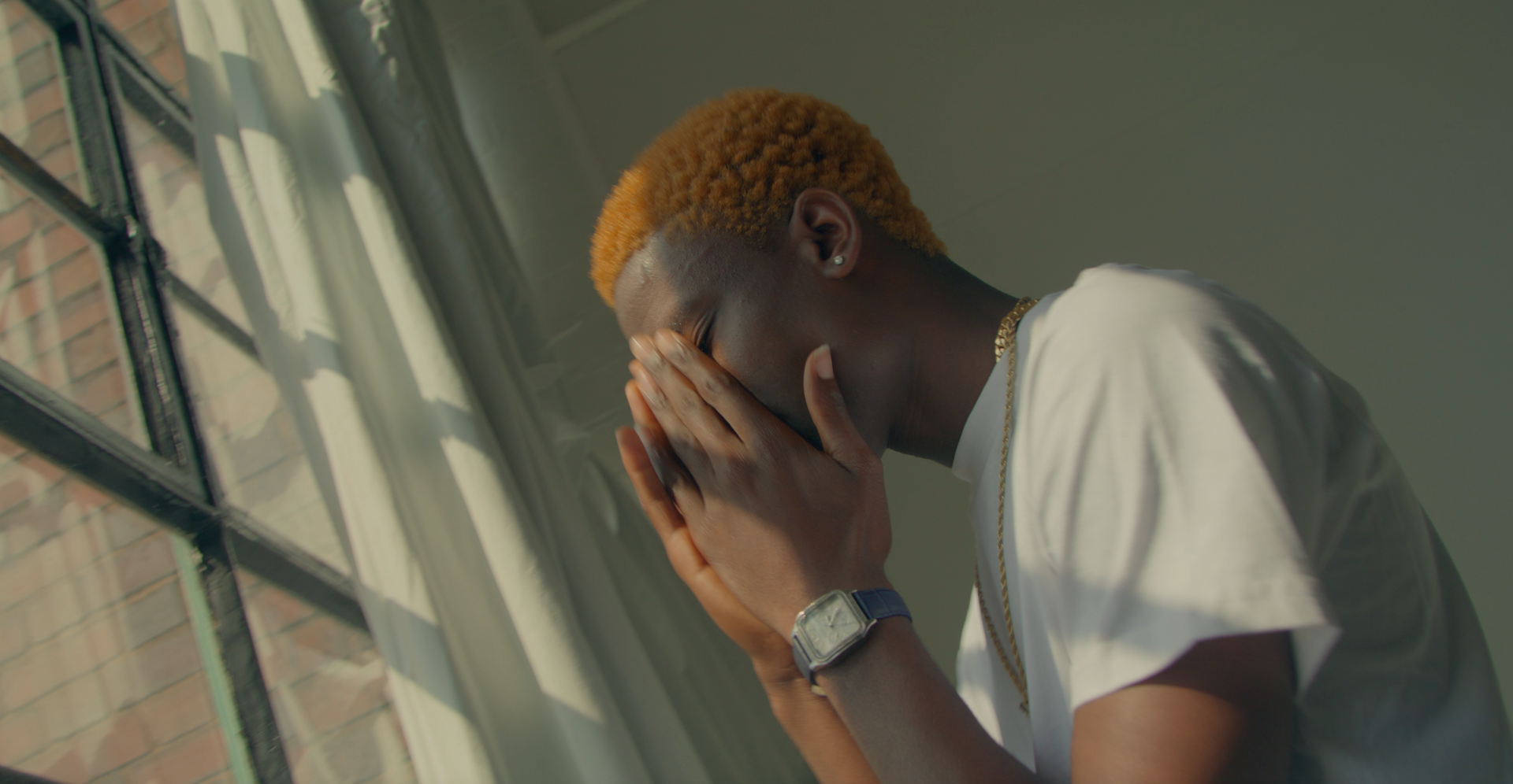 A man with red hair is sitting in front of a window covering his face with his hands.