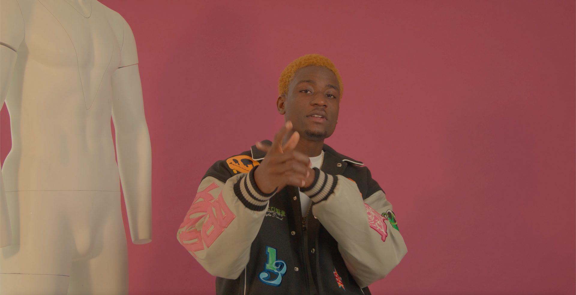 A man in a jacket is pointing at a mannequin on a pink background.
