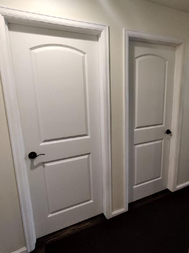 Two white doors with arched tops and black handles in a hallway with white trim and dark flooring.