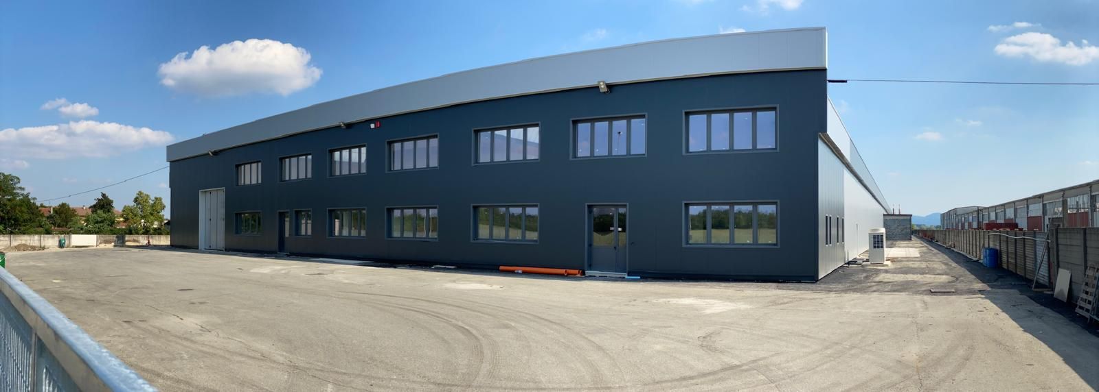 Edificio industriale grigio scuro con numerose finestre che si affaccia su un lotto pavimentato sotto un cielo azzurro.