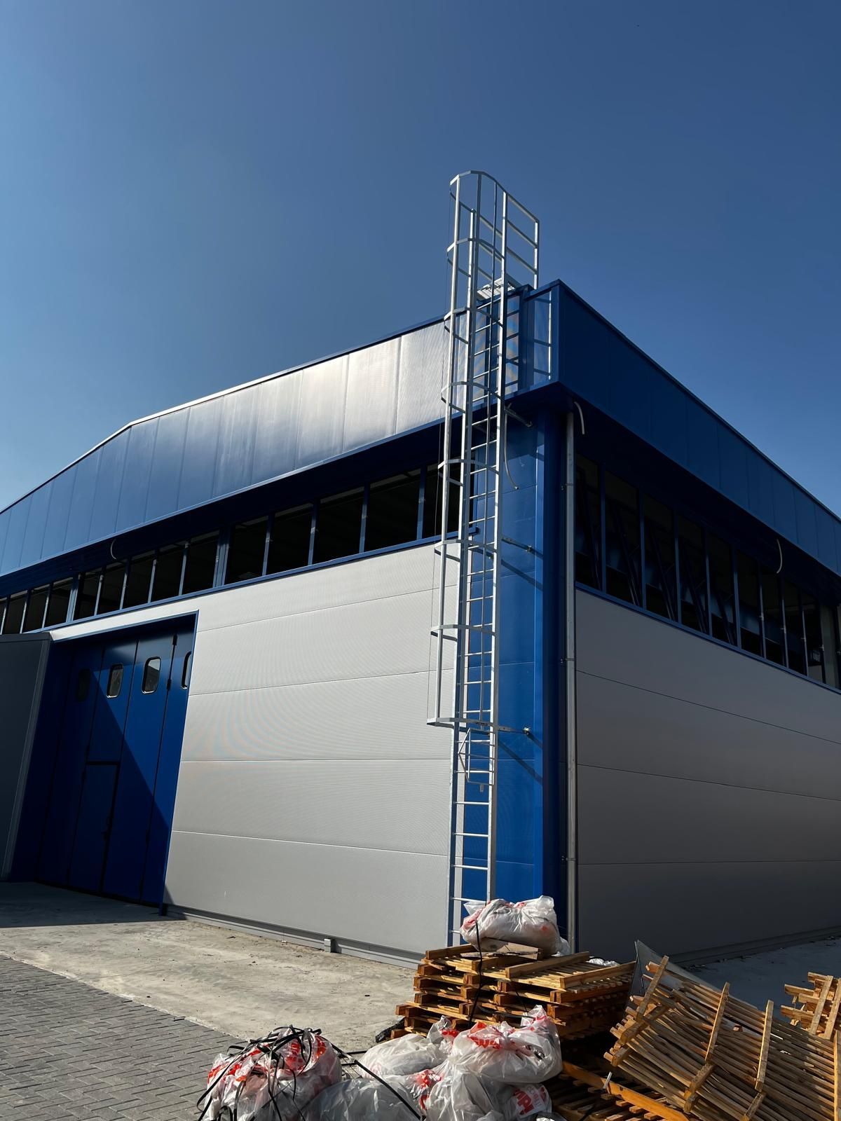 Edificio industriale blu con rivestimento metallico e impalcature, con pallet impilati e detriti all'esterno.
