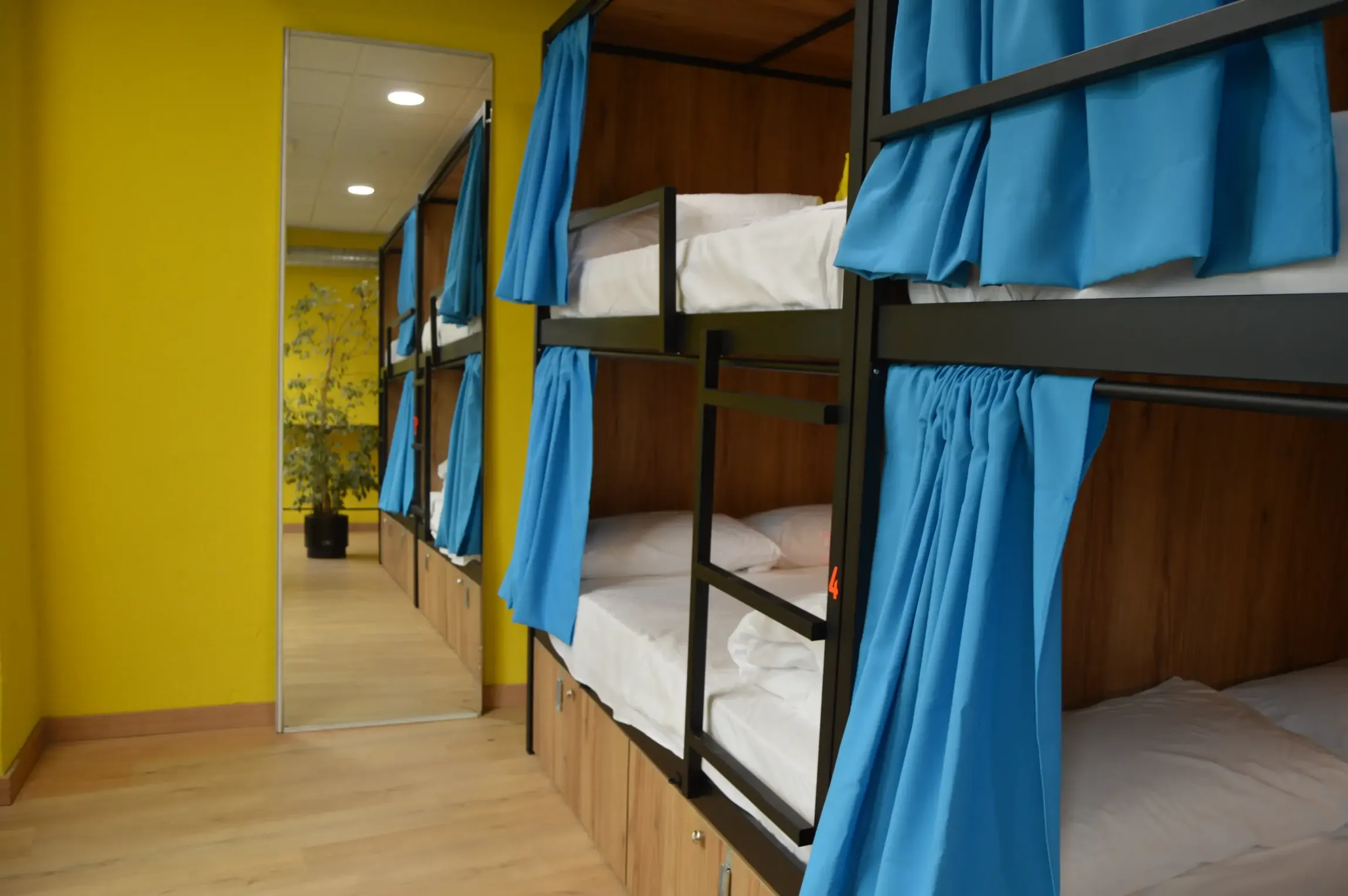 Literas en una habitación muy iluminada con paredes amarillas y cortinas azules. Hay un espejo largo en la pared.
