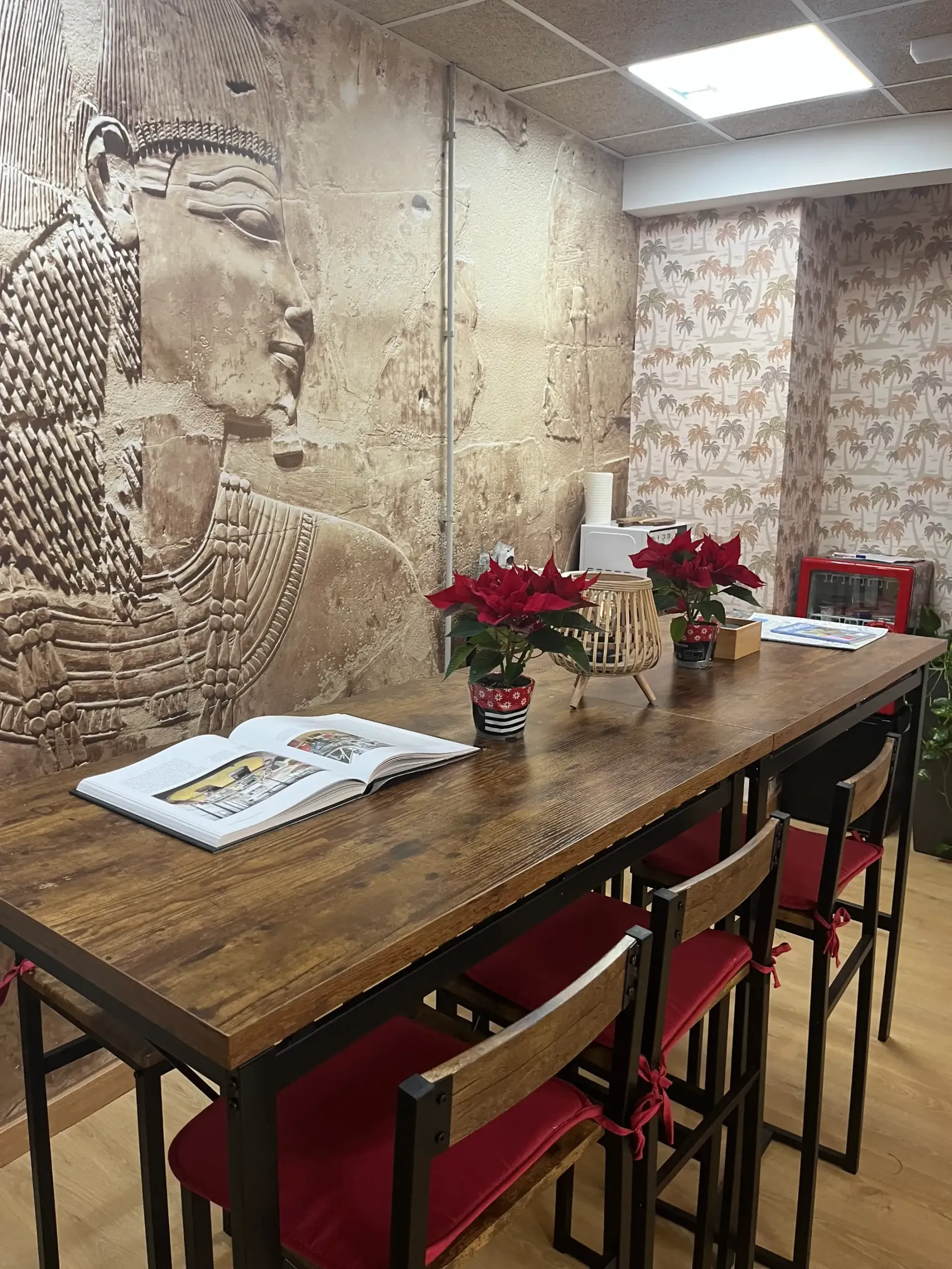 Una larga mesa de madera con taburetes de cojines rojos, bajo un mural de temática egipcia y flores de Pascua rojas.