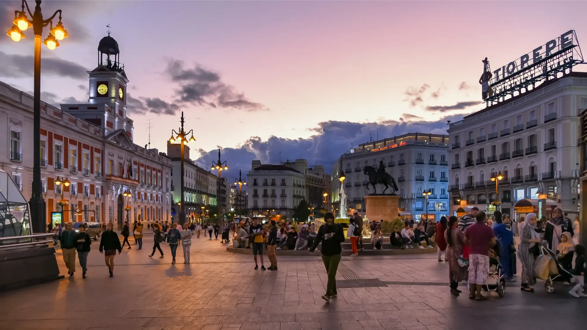 Plaza del Sol, Madrid, al atardecer. Se ven edificios, farolas y una estatua mientras la gente pasea. Cielo rosa y morado.