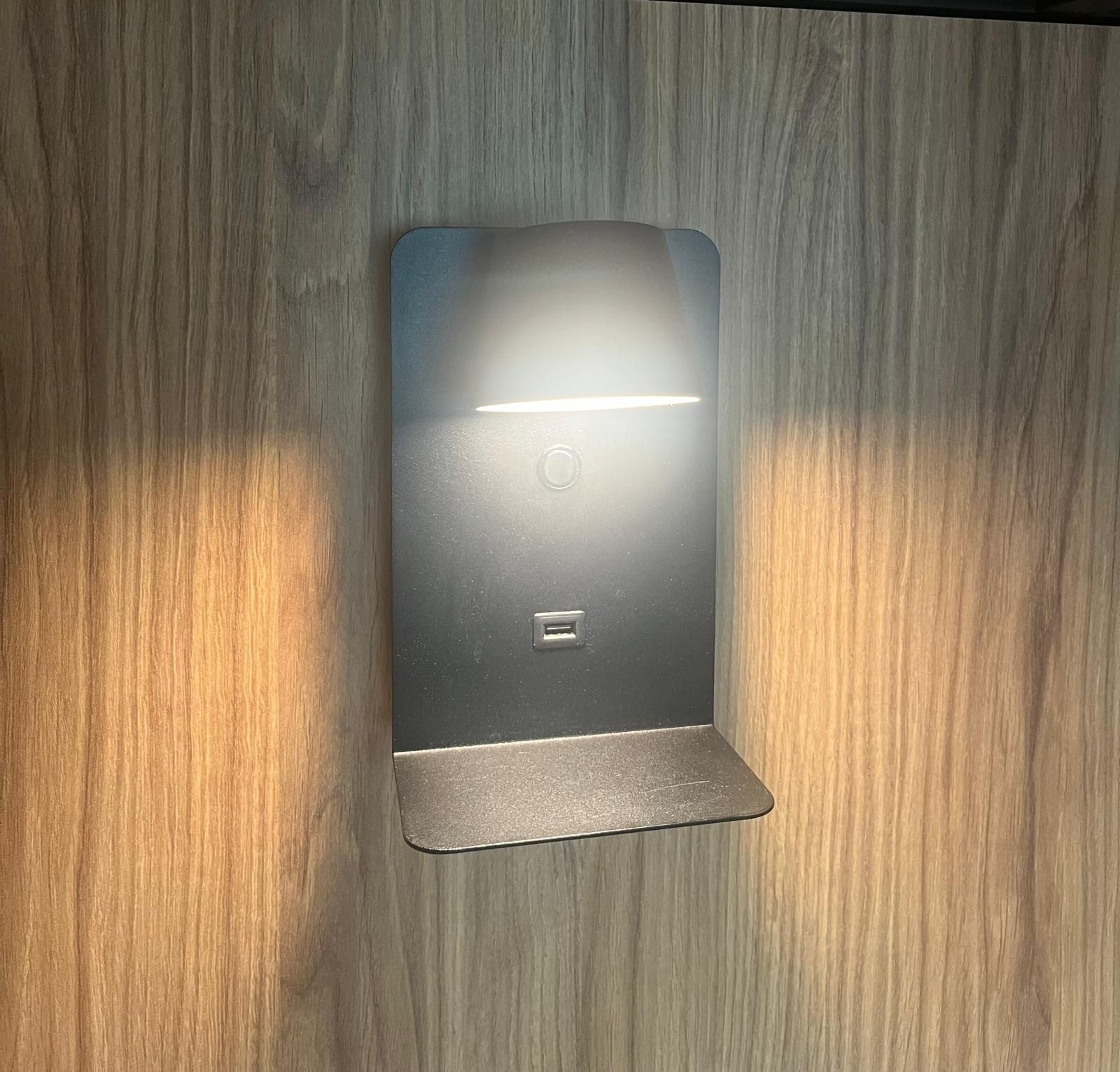 Aplique de pared rectangular negro con barra de luz horizontal y pequeño puerto de carga sobre superficie de madera.