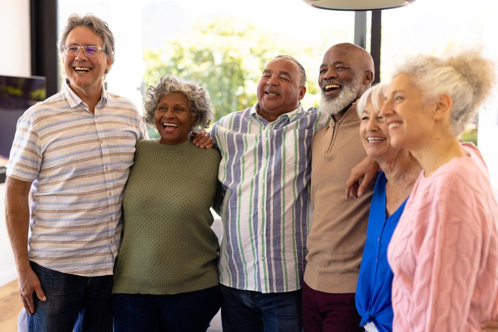 Group of friends, arms around each other, laughing together in a well-lit room with large windows.