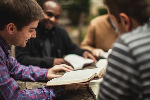 Four people smiling, reading books around an outdoor table.