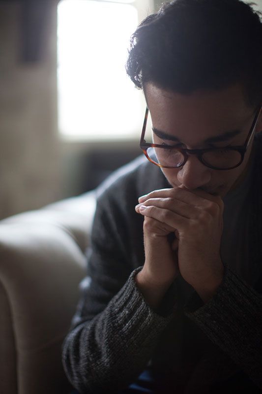 Man with glasses, hands clasped, head bowed, in thought. Soft light from a window.
