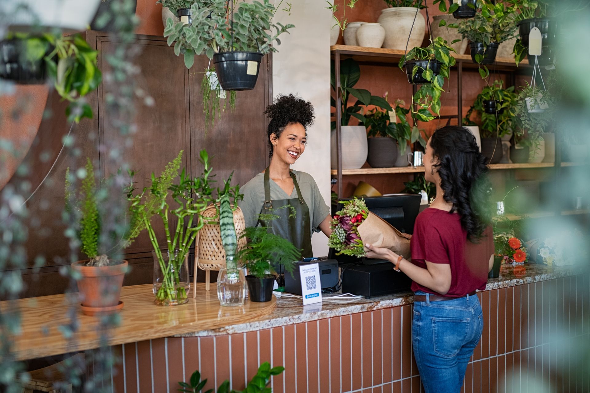 Two people at a plant shop counter, one handing over a potted plant amid hanging greenery.