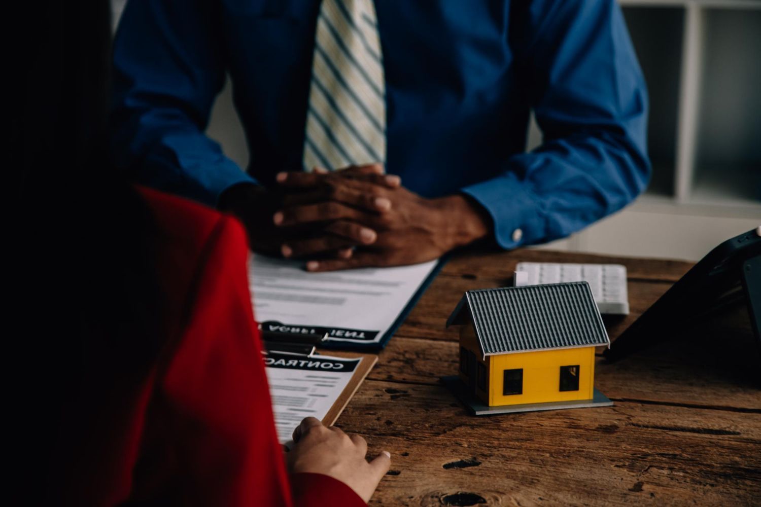 Two people discuss paperwork at a table with a small yellow house model in the foreground.