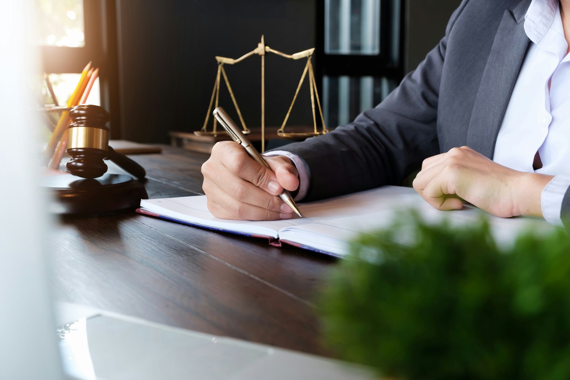 Lawyer writing at a desk beside a brass scales of justice in a dim office