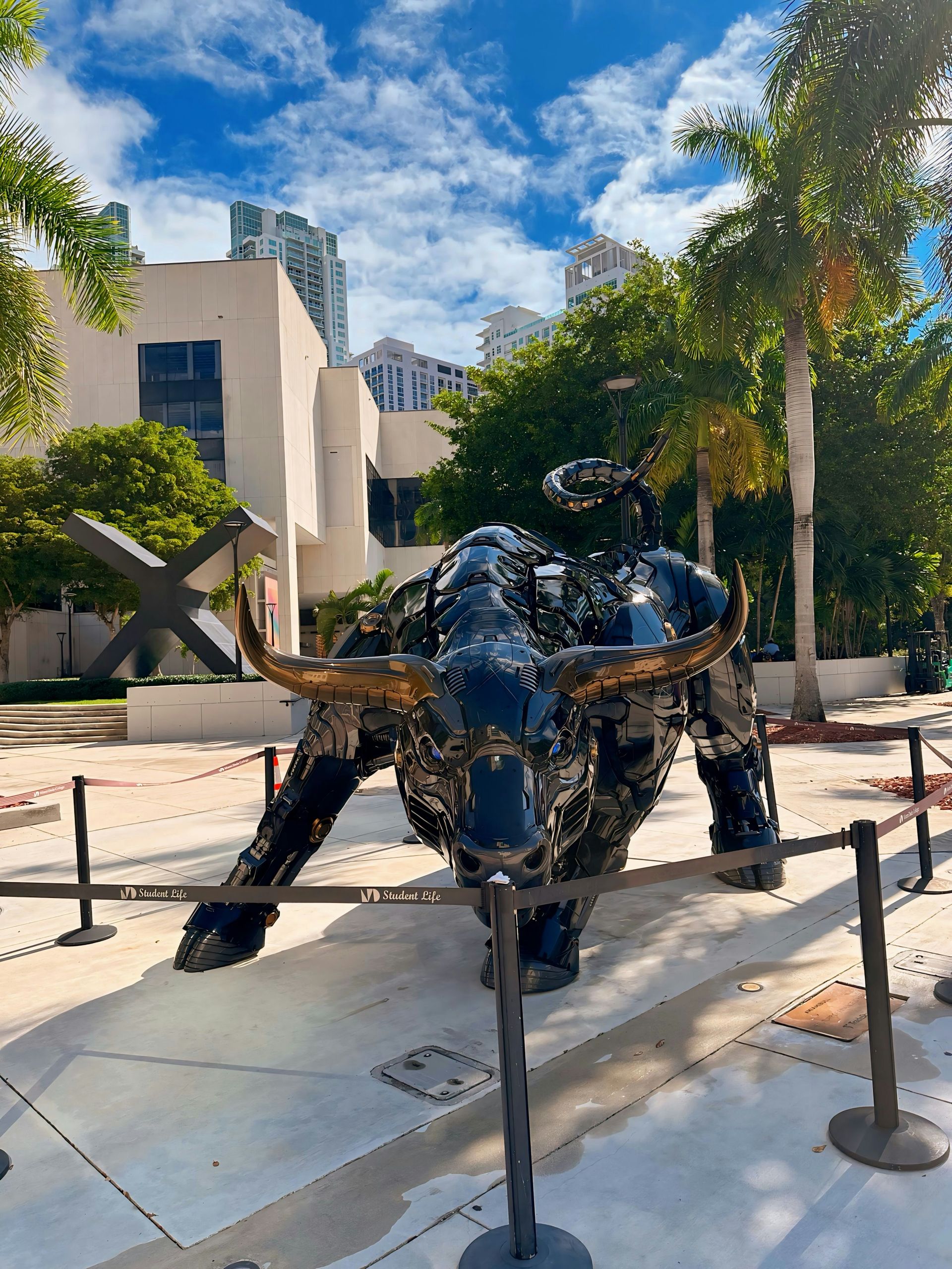 A black bull sculpture in a plaza, surrounded by barriers, with buildings and palm trees under a blue sky.