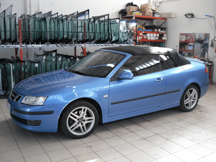 Auto decappottabile blu all'interno di un'autofficina con file di parabrezza sullo sfondo.