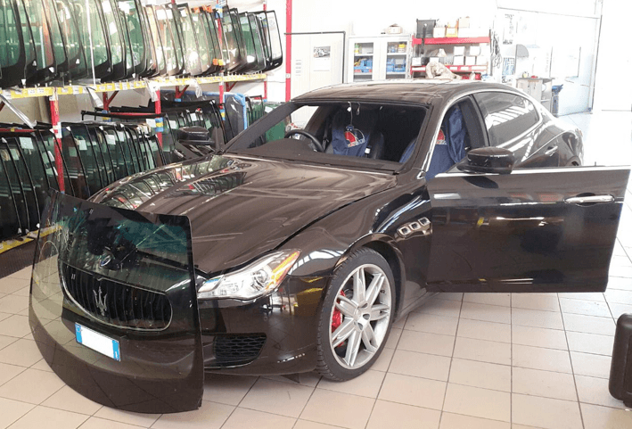 Berlina Maserati nera con portiera aperta, parabrezza coperto, in un'officina di riparazione vetri.