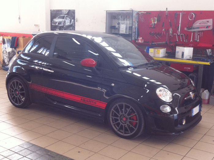 Fiat 500 Abarth nera con dettagli rossi e strisce da corsa, parcheggiata all'interno di un garage.