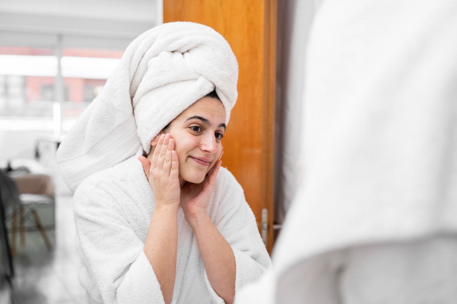 A woman with a towel wrapped around her head is looking at her face in the mirror.