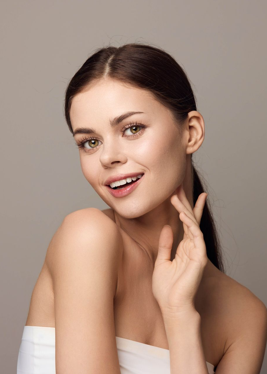 Woman with dark hair smiles, touches her neck, and looks at the viewer; neutral background.