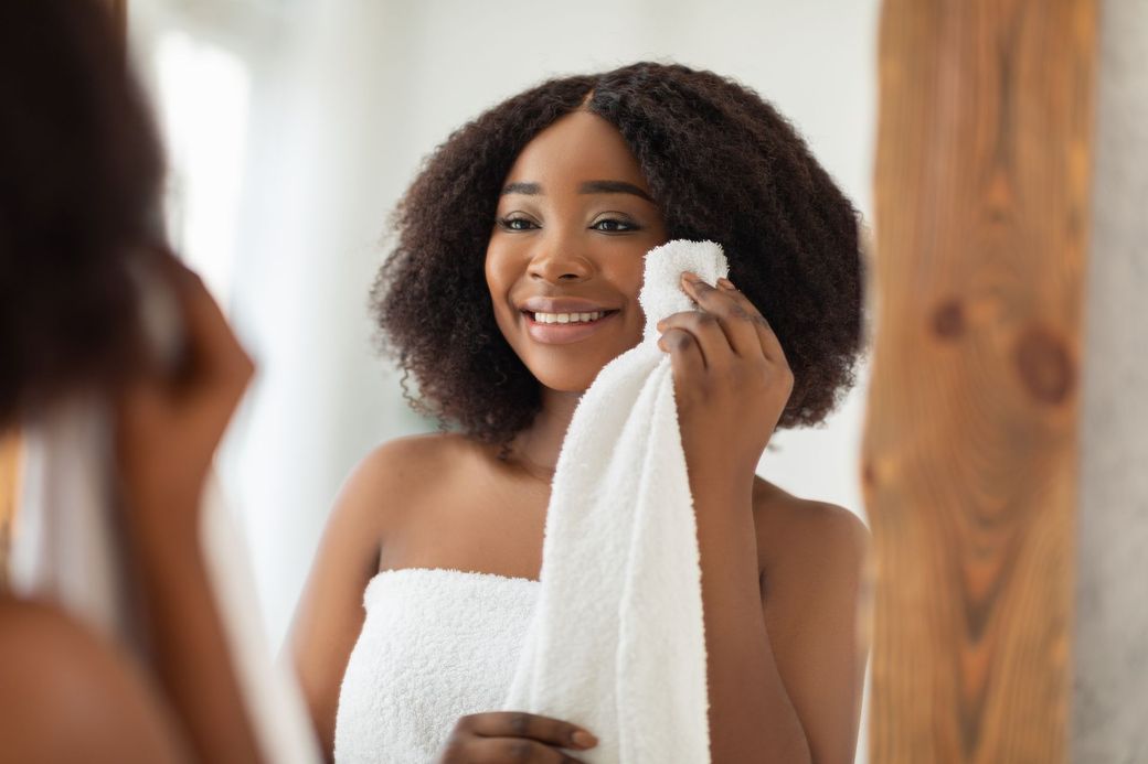 A woman wrapped in a towel is cleaning her face in front of a mirror.